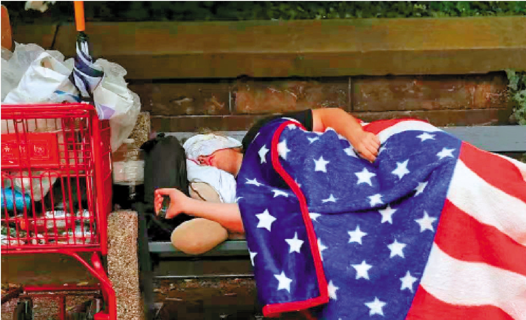 　　圖：美國民眾一旦跌破「斬殺線」，就可能遭遇生存危機，甚至流落街頭。圖為一名美國無家可歸者披着國旗睡覺，以公園為家。