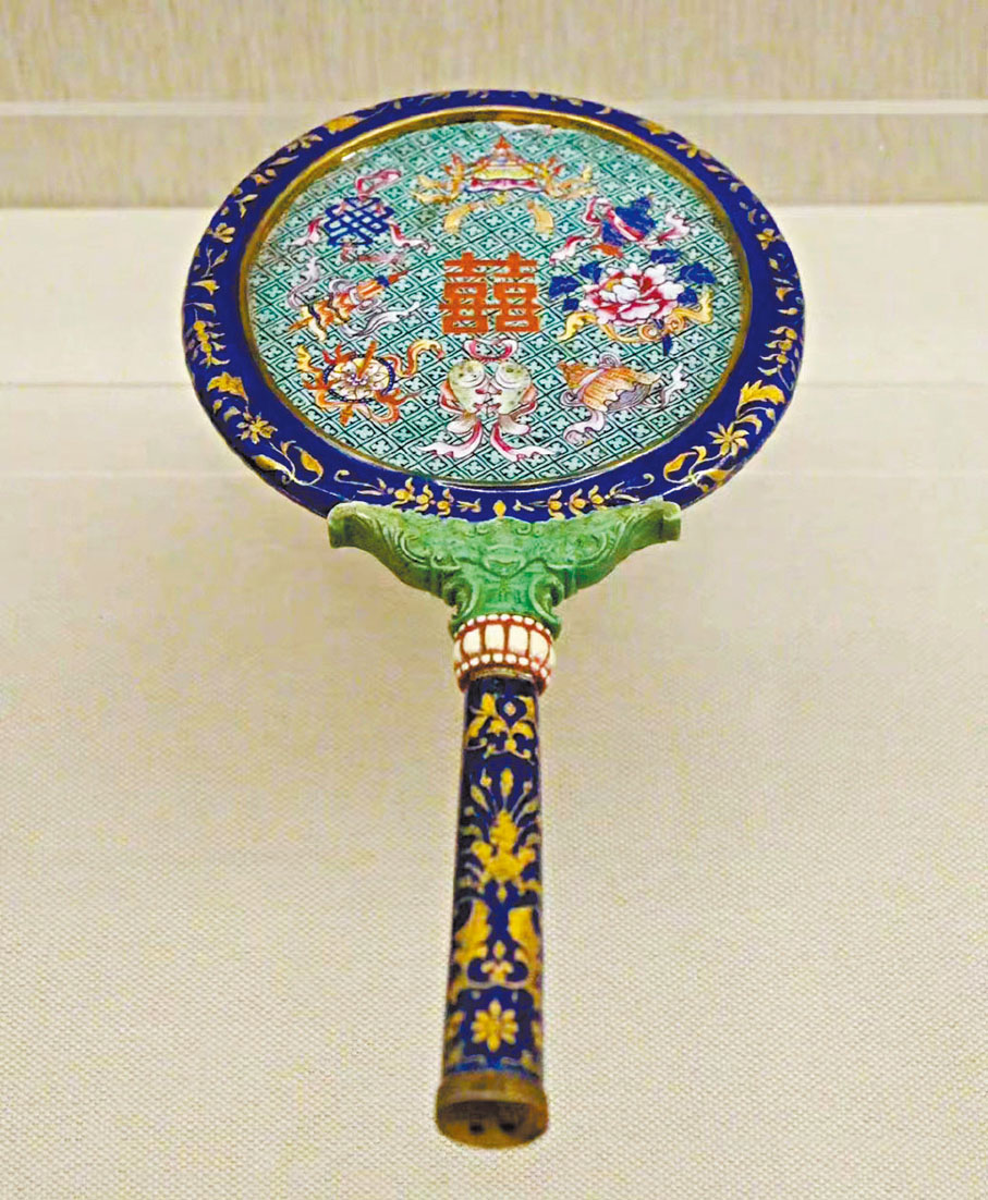 ◆清代畫琺瑯八寶喜字紋把鏡。鏡身呈橢圓形，正面為水銀玻璃鏡，背面以「卍」字紋為底，正中畫雙喜字，四周環繞八寶紋，外邊鑲嵌藍色琺瑯描金花卉紋鏡圈。