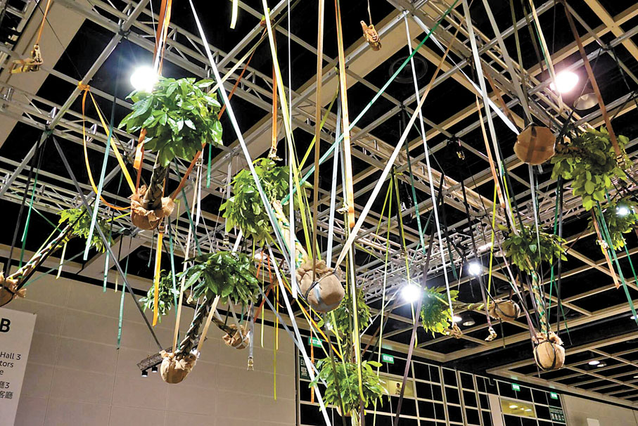 【楊沛鏗《屋簷下的擁抱先生們》】《屋簷下的擁抱先生們》由13棵懸掛的發財樹組成，以強壯堅實和吉祥意味見稱的發財樹在作品中相互交織的狀態象徵了生命間的彼此糾纏。這件作品呈現出在被隔離和惴惴不安之中人與人的互相依賴，展現出一種集體的緊張和無力感，也象徵了團結和韌力。