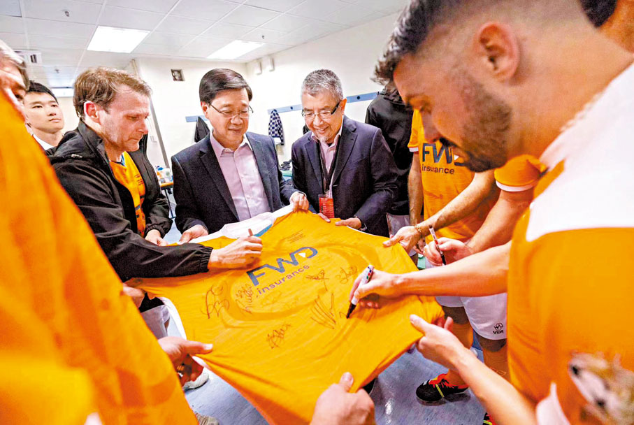 ◆特首李家超在球員更衣室和世界明星隊的球員互動，還獲贈送球衣，球員正在球衣上簽名。