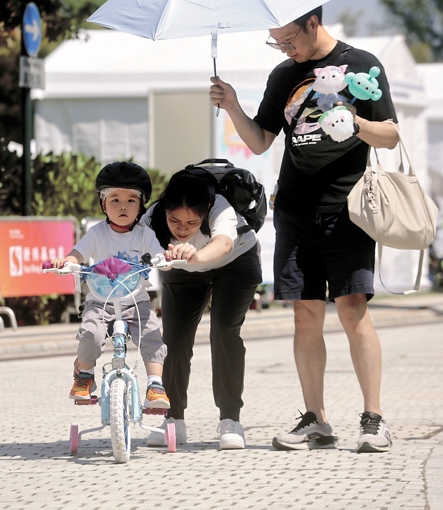 ●爸爸為妻子女兒撐着傘遮擋太陽，媽媽從後抓緊單車，讓女兒體驗騎單車的感覺，一家共享天倫之樂。